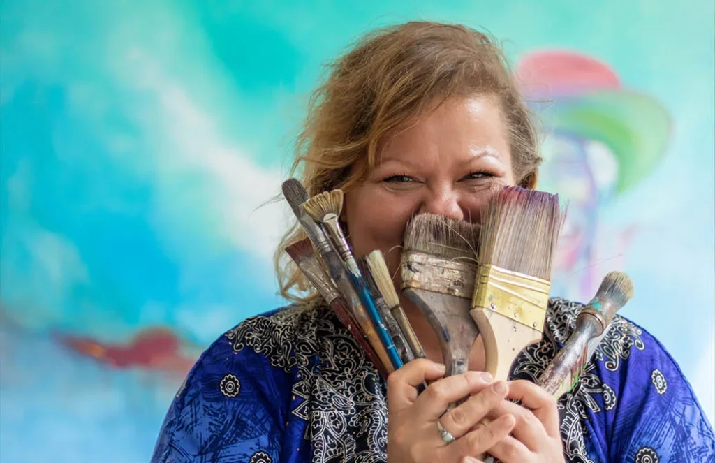 smiling woman holding up paint brushes to her face