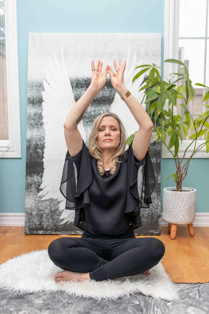 Woman seated cross‑legged with raised arms in front of angel wings artwork, serene lifestyle branding portrait.