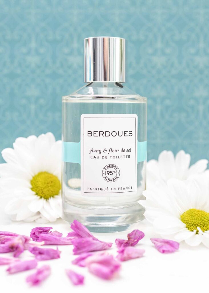 Editorial product photo of Berdoues Ylang & Fleur de Sel perfume bottle styled with flowers on a blue textured background.