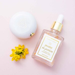 Flat‑lay product photo of Fancy Face Poetry in Potion facial serum with gold dropper, white compact, and yellow flowers on a soft pink background.
