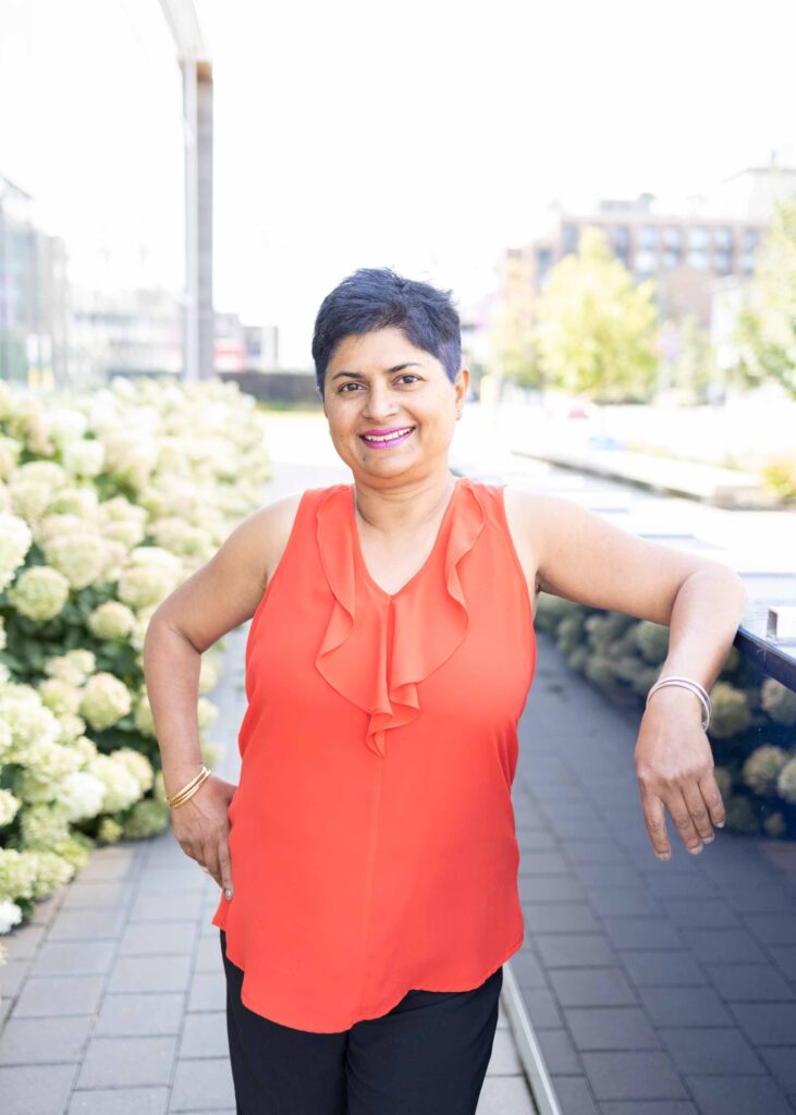 Personal branding portrait of Cheryl Pais standing outdoors in a coral top, smiling beside greenery — photographed by Marta Raptis in Oakville.