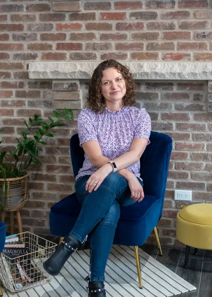 Marta Raptis, brand photographer, seated on a navy velvet chair in a cozy studio with brick wall and plants.