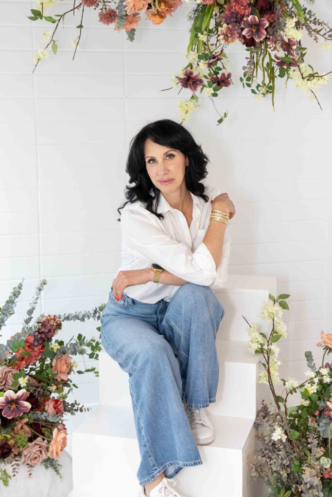 Allison Jorgens seated in white shirt and jeans with floral backdrop, photographed for Qwiky Wellness branding shoot by Marta Raptis.