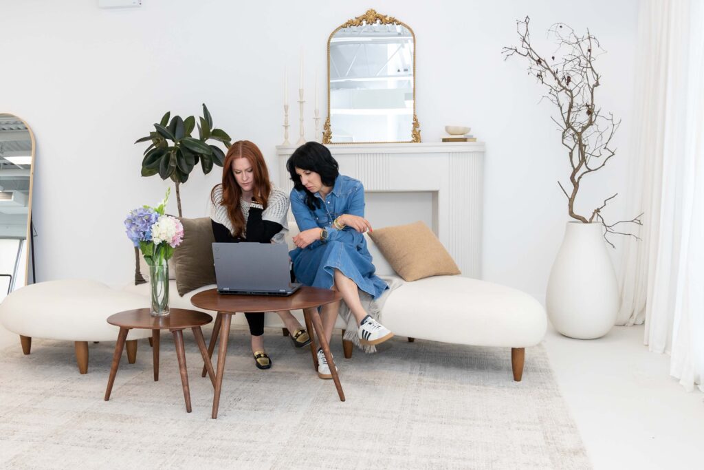 Samara Foisy and Allison Jorgens collaborating on laptops in bright studio with neutral decor, photographed for Qwiky Wellness branding shoot by Marta Raptis.