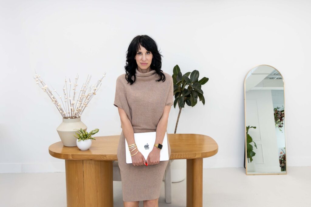 Samara Foisy standing with laptop and notebook in bright studio with plants, photographed for Qwiky Wellness branding shoot by Marta Raptis.