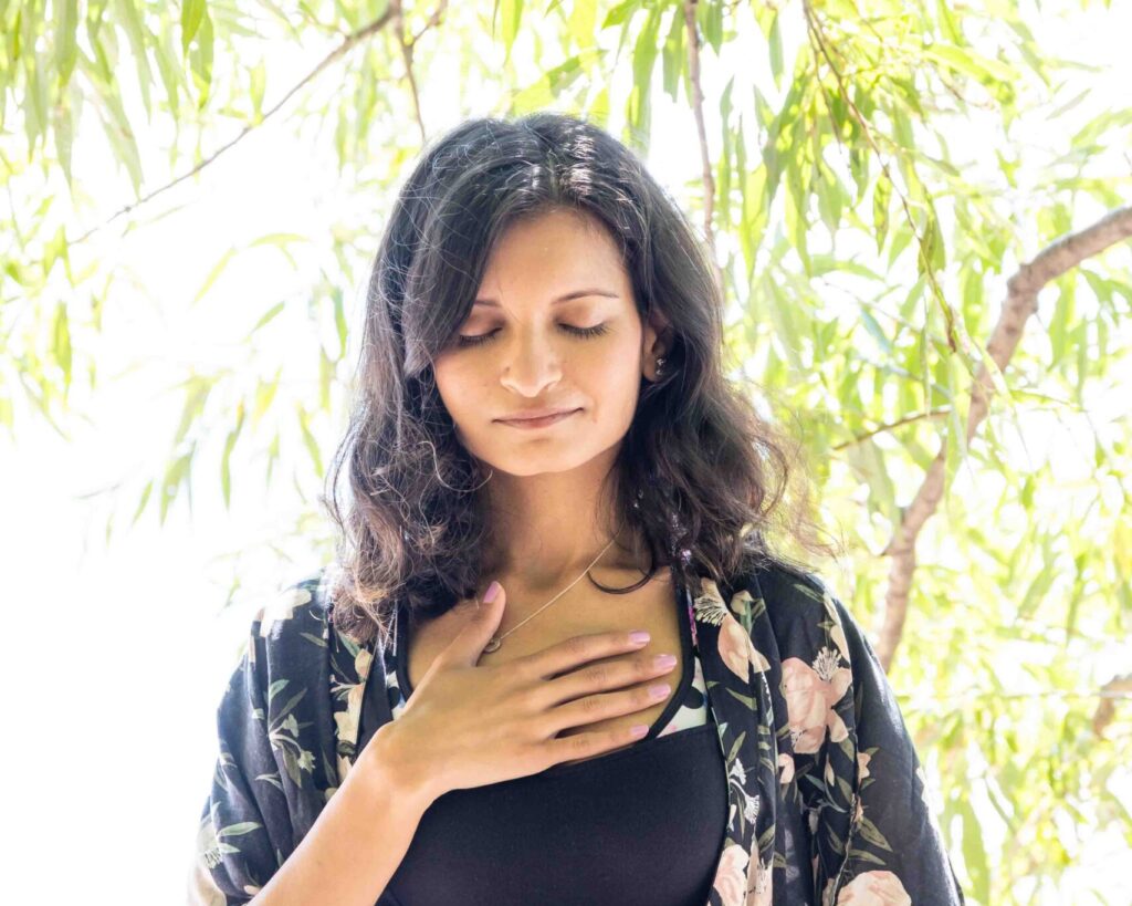 Woman standing outdoors with hand on chest, wearing a floral kimono, surrounded by soft natural light and greenery.
